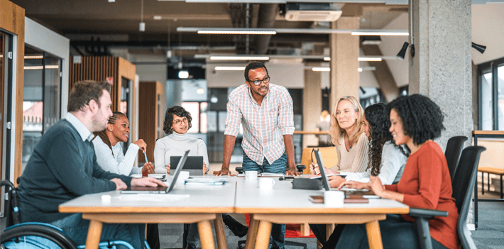Bright open concept office. There is a work meeting being held where a group of diverse people, including a man on a wheelchair are working together.