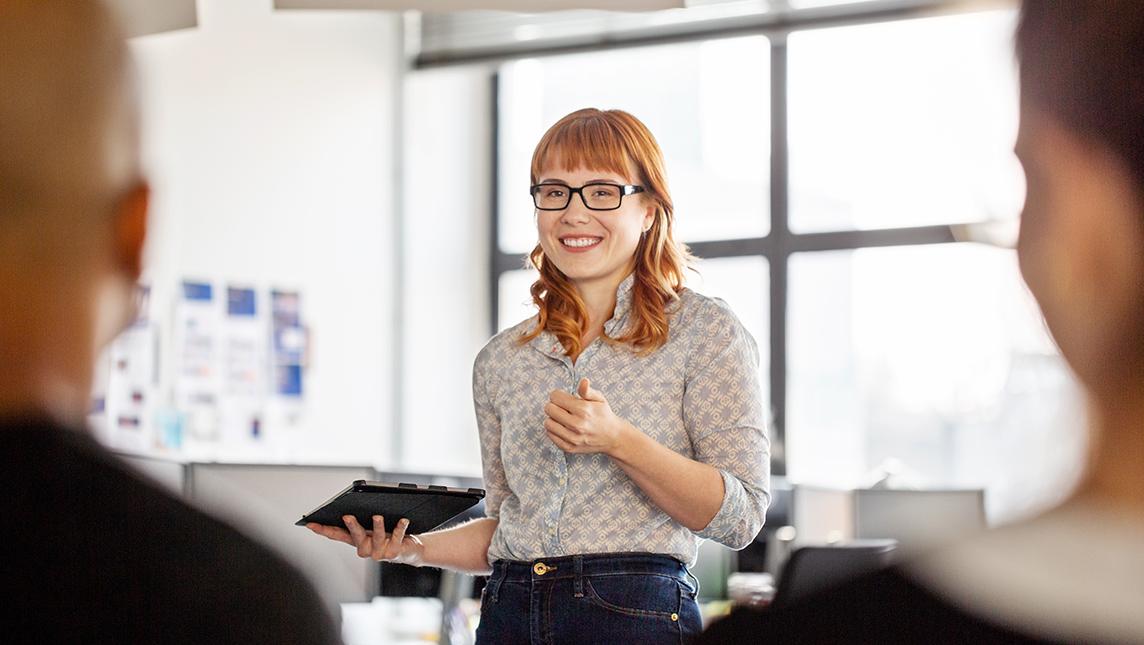 A woman with a tablet addresses an audience of work colleagues.