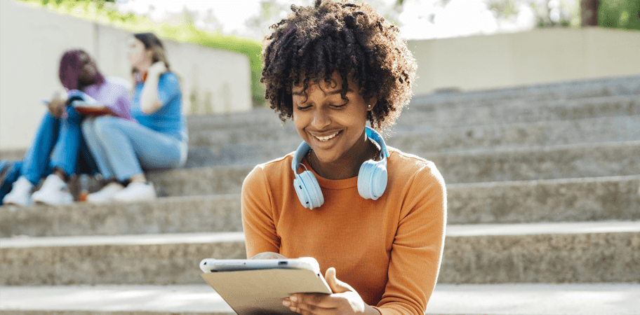 Latin young woman at school recess sitting on some stairs in the park with blue headphones around her neck and looking at a digital tablet while smiling.