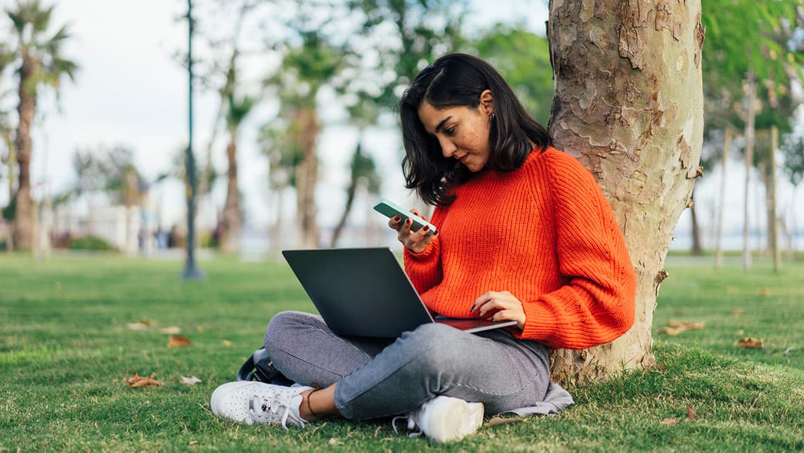 A student sits under a tree using her laptop and phone.