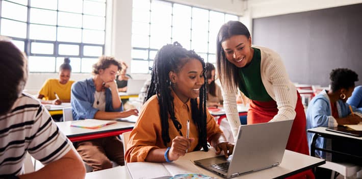 Smiling teacher talking with a student using a laptop during a classroom lesson