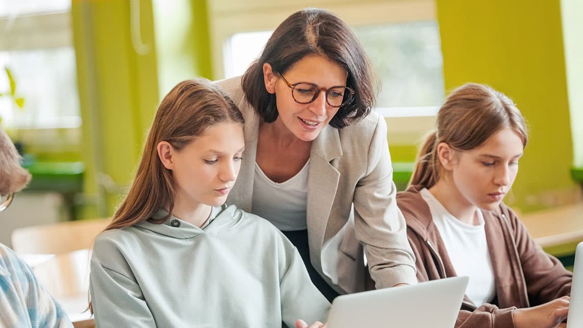 A teacher helps students on their laptops in a classroom.