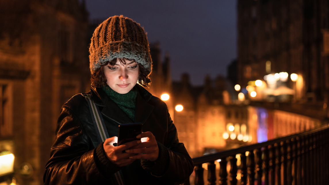 A woman using her phone on a city in Edinburgh's Old Town at dusk in winter.