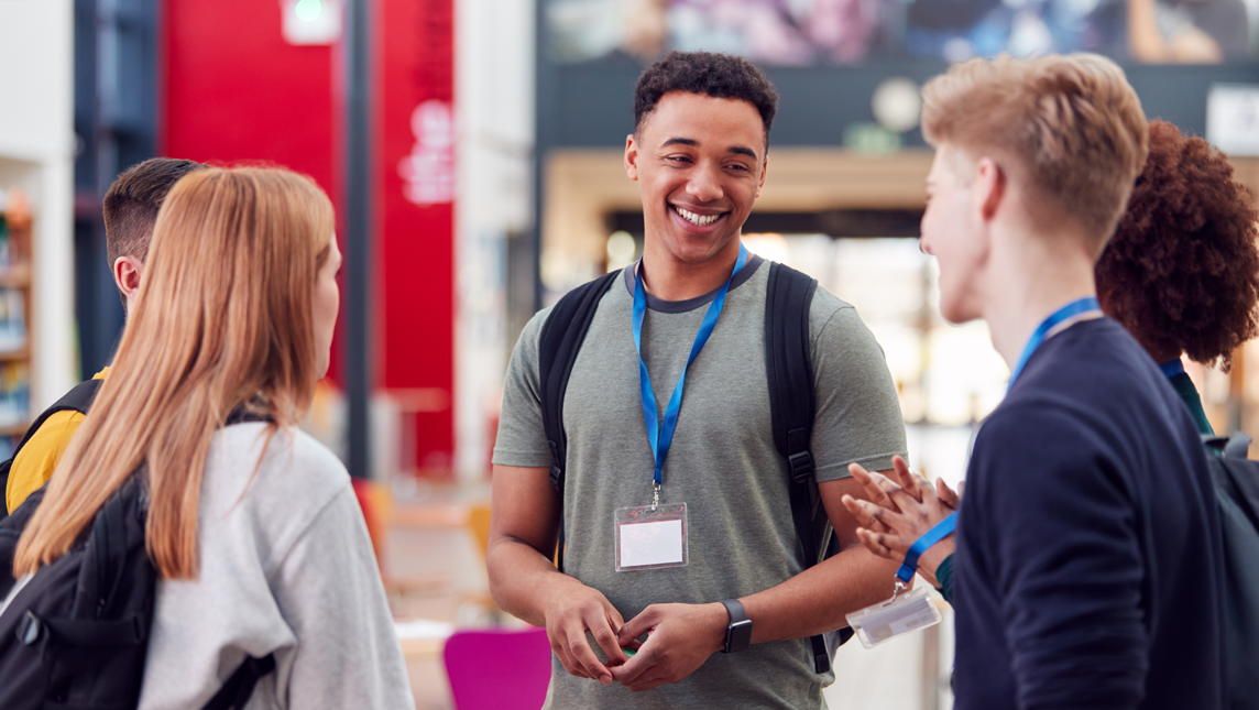 A group of five young adults are standing in a circle, engaged in conversation. They are wearing casual clothing and have lanyards with name tags around their necks.