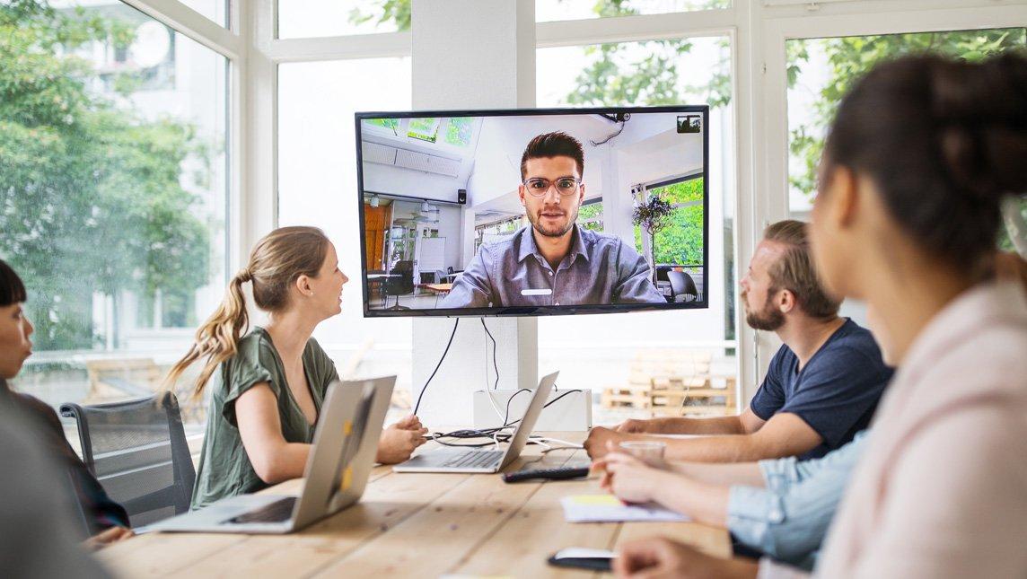 Four people seated around a wooden table in a bright, modern office, participating in a video conference call displayed on a large wall-mounted screen.
