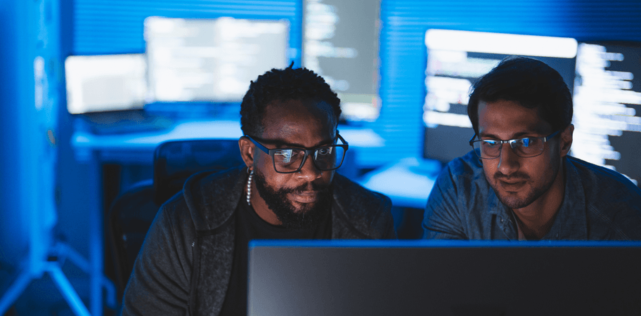 Two people working together at a computer in a dimly lit room with multiple monitors displaying code and technical information in the background.